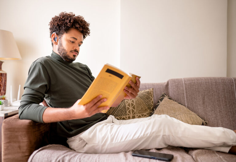 Adult male with a hearing aid reading a book while relaxing on his living room sofa at home