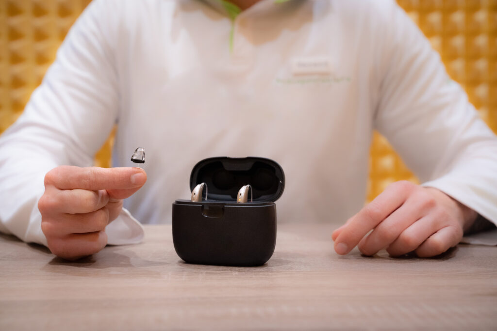 rechargeable hearing aids - Doctor audiologist in their office with one hearing aid in his hand and two hearing aids in their case. Showing care tips.